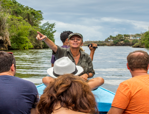 Guide sitting with people on a boat facing the camera and pointing at something in front of her