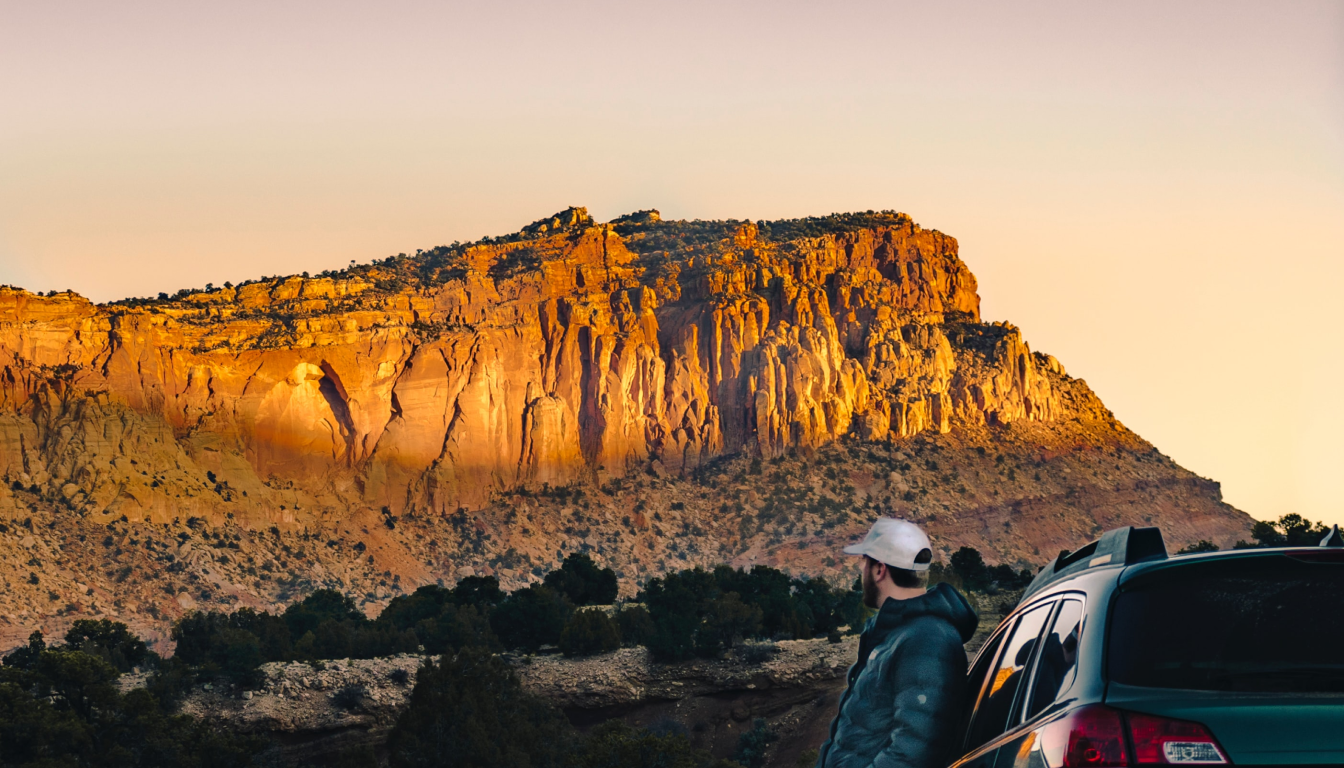 Man standing with his back against his car looking at a rocky hill at sunset